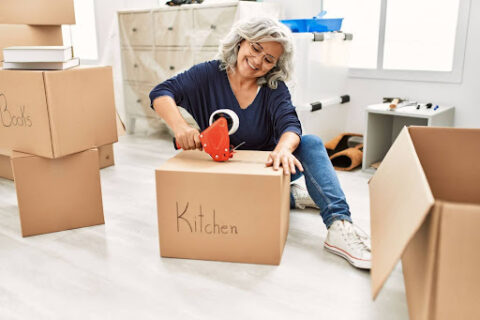A senior woman sits on the floor surrounded by moving boxes and smiles as she puts packing tape on a moving box marked “kitchen.”