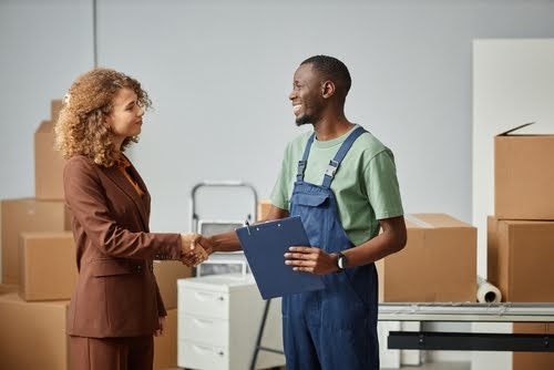 Young businesswoman concluding a deal with worker from moving service, they standing in office and shaking hands to each other